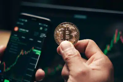 Una mano sostiene una moneda física de Bitcoin frente a un smartphone que muestra gráficos de trading en tiempo real.