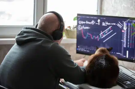 Una persona de espaldas con audífonos puestos, concentrada frente a un monitor que muestra gráficos de trading y velas japonesas.