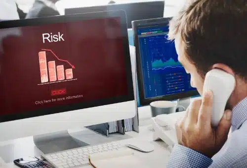 Un hombre hablando por teléfono mientras observa dos monitores. La pantalla principal muestra un fondo rojo con la palabra "Risk" (Riesgo) y un gráfico de barras que indica una caída drástica