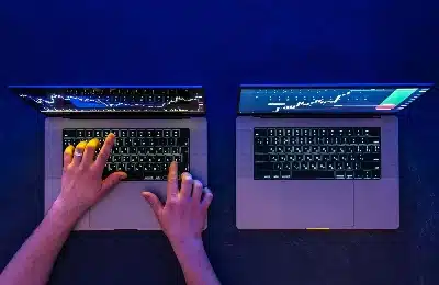 vista aérea de dos computadoras portátiles abiertas sobre una superficie oscura bajo una iluminación azul intensa. En las pantallas se aprecian gráficos de velas japonesas y análisis de mercados financieros