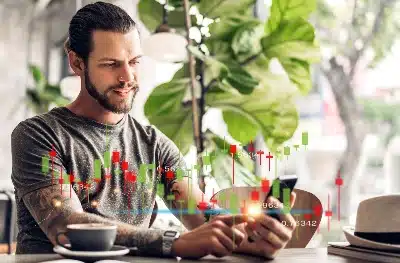 Un hombre con tatuajes observa con interés su teléfono móvil en un café, mientras sobre la pantalla se superpone una interfaz digital de velas japonesas y datos financieros en tiempo real.