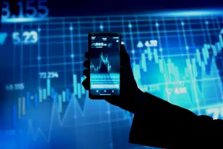 mano sosteniendo un smartphone frente a una pantalla gigante con gráficos financieros en tonos azules. En la pantalla del celular también se aprecia un gráfico de velas en tiempo real, lo que sugiere el uso de una aplicación de trading o seguimiento de criptomonedas.