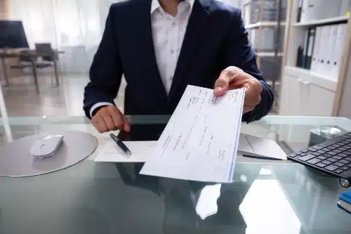 Un hombre de negocios sentado tras un escritorio de cristal, extendiendo la mano para entregar un cheque bancario en primer plano.