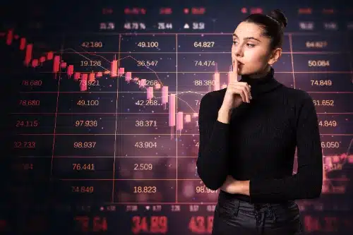Una mujer pensativa con un dedo en los labios, frente a una gran pantalla que muestra gráficos financieros con una tendencia a la baja.
