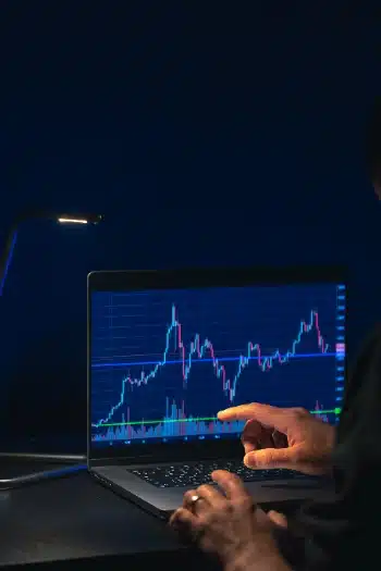 una persona analizando gráficos de trading en una laptop durante la noche. La pantalla muestra fluctuaciones del mercado con velas japonesas y líneas de tendencia sobre un fondo oscuro, mientras una lámpara de escritorio ilumina suavemente las manos del usuario