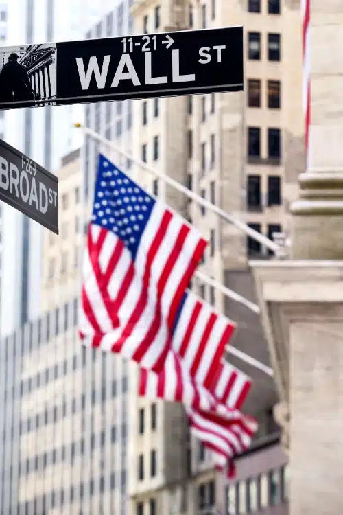 Un cartel callejero de Wall St y Broad St en primer plano, con varias banderas de Estados Unidos y edificios financieros desenfocados al fondo.