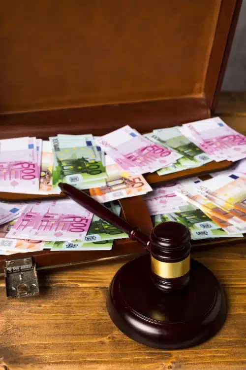 Primer plano en plano vertical de un mazo de juez de madera oscura apoyado sobre una mesa frente a un maletín de cuero marrón abierto y repleto de billetes de euro de varias denominaciones