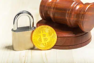una moneda dorada de Bitcoin se encuentra en el centro, flanqueada por un candado metálico cerrado a la izquierda y un mazo de juez de madera con su base a la derecha