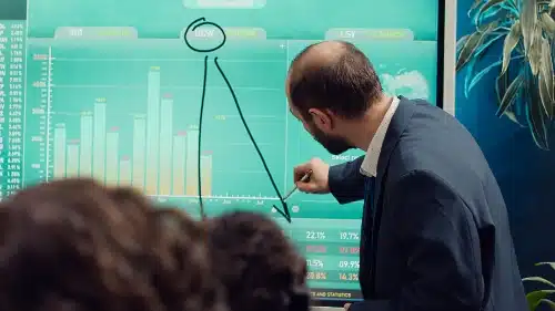 se muestra de perfil a un hombre con traje que utiliza un bolígrafo digital para dibujar una línea descendente en una gran pantalla interactiva verde, la cual despliega gráficos y datos de un analizador de acciones frente a un equipo.