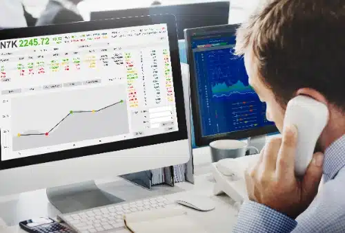 se observa de espaldas a un hombre hablando por teléfono mientras analiza gráficos financieros y tablas de datos bursátiles en dos monitores en su escritorio de oficina.