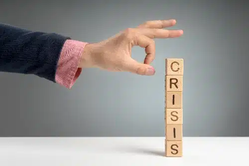 una mano con traje oscuro y camisa rosa está a punto de empujar o tocar una torre vertical de bloques de madera que forman la palabra "CRISIS", sobre un fondo gris.