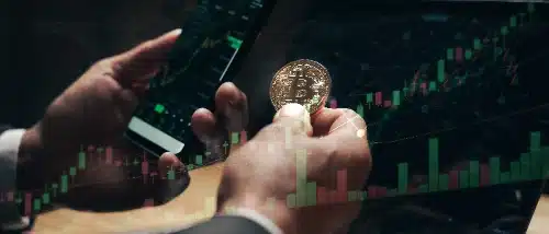 Una persona sostiene una moneda dorada de Bitcoin mientras revisa un smartphone, con un gráfico bursátil superpuesto.