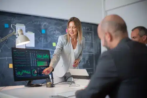 Una líder empresarial en traje apunta hacia un monitor con gráficos financieros de rendimiento y paneles de datos mientras explica la información a dos colegas.