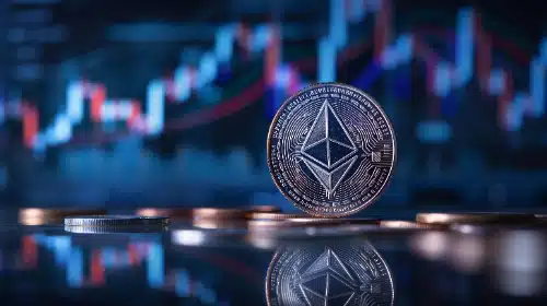 Una moneda física de Ethereum se mantiene de pie sobre una superficie reflectante rodeada de otras monedas. Al fondo, se muestra un gráfico financiero de velas de trading borroso en tonos azules y rojos.
