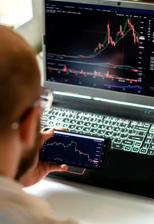 Un hombre sostiene un teléfono móvil en su mano mostrando un gráfico de líneas financieras frente a una laptop.