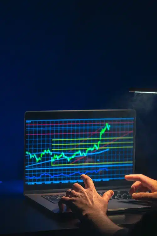 Un primer plano de las manos de una persona operando en una computadora portátil que muestra gráficos financieros con líneas verdes y cuadrículas.