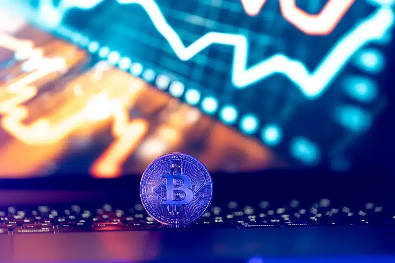 Una moneda de Bitcoin iluminada con tonos morados descansa sobre el teclado de un portátil, con un gráfico de líneas neón brillante al fondo.