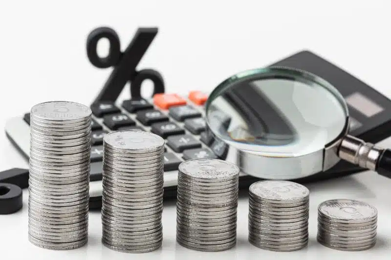 Pilas de monedas ordenadas de mayor a menor altura frente a una calculadora, una lupa y un símbolo de porcentaje sobre un fondo blanco.