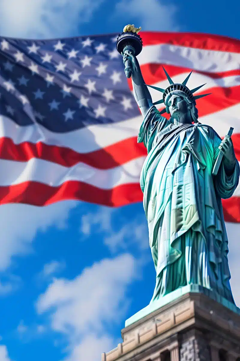 La Estatua de la Libertad se yergue majestuosa sobre su pedestal con la antorcha en alto, respaldada por una gran bandera estadounidense ondeando bajo un cielo azul con nubes.