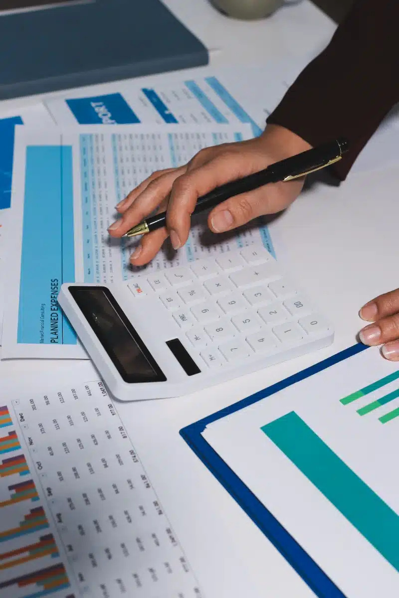 Una persona utiliza una calculadora blanca y sostiene un bolígrafo para revisar informes de gastos y gráficos financieros sobre el escritorio.