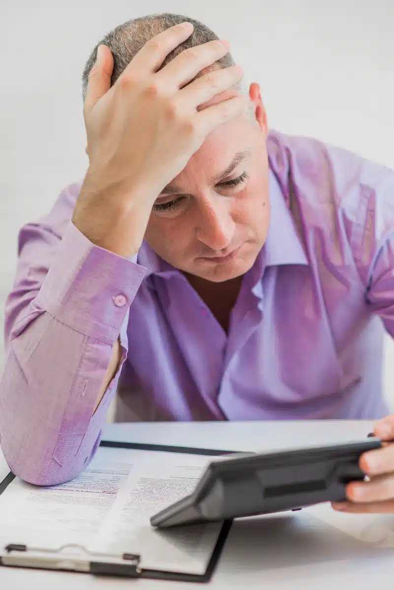 Hombre preocupado sosteniendo su cabeza mientras revisa documentos financieros y usa una calculadora sobre su escritorio.