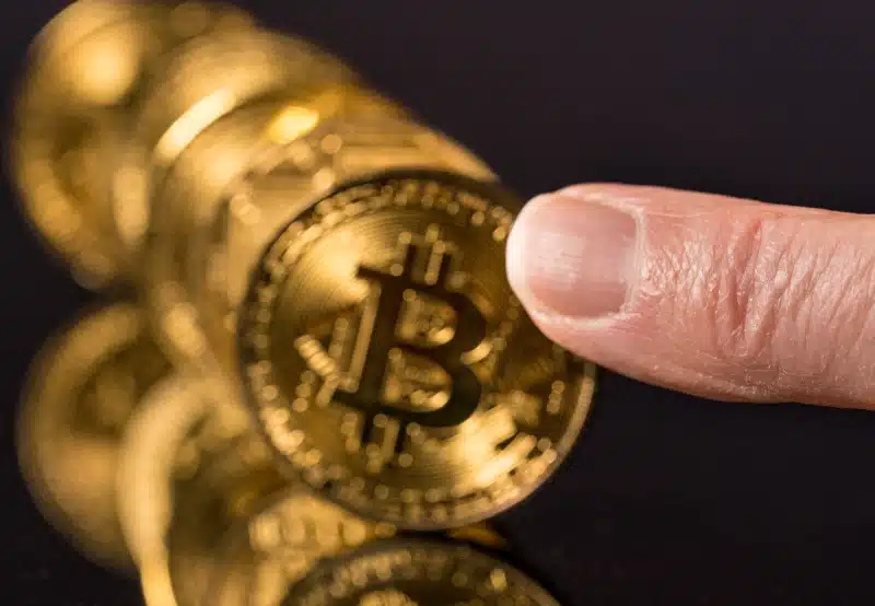 Dedo apunto de empujar una hilera de monedas doradas de Bitcoin alineadas como fichas de dominó, simbolizando la reacción en cadena y el riesgo en el mercado de criptomonedas.