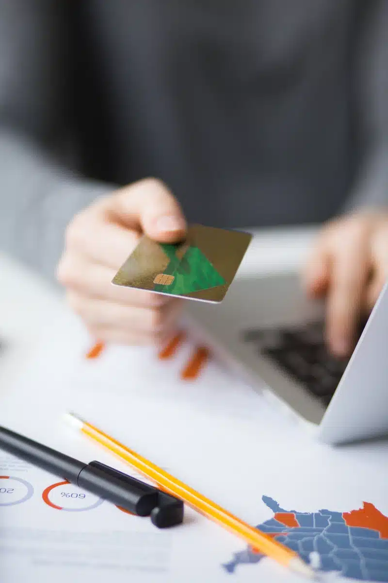 Persona sosteniendo una tarjeta de crédito dorada frente a una computadora portátil sobre un escritorio con documentos financieros y material de oficina, representando transacciones electrónicas y banca en línea.