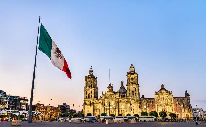 Bandera monumental de México ondeando en el Zócalo con la Catedral Metropolitana al atardecer en Ciudad de México.